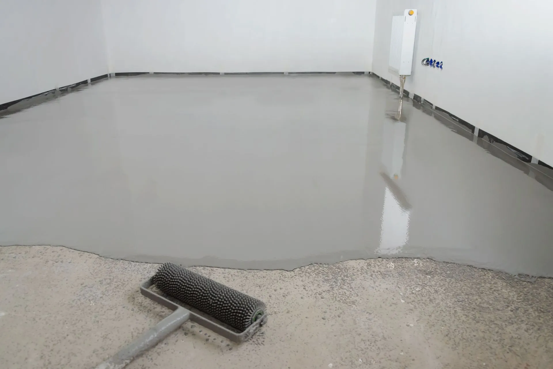 Spiked roller spreading a fresh gray self-leveling coat across a commercial concrete floor